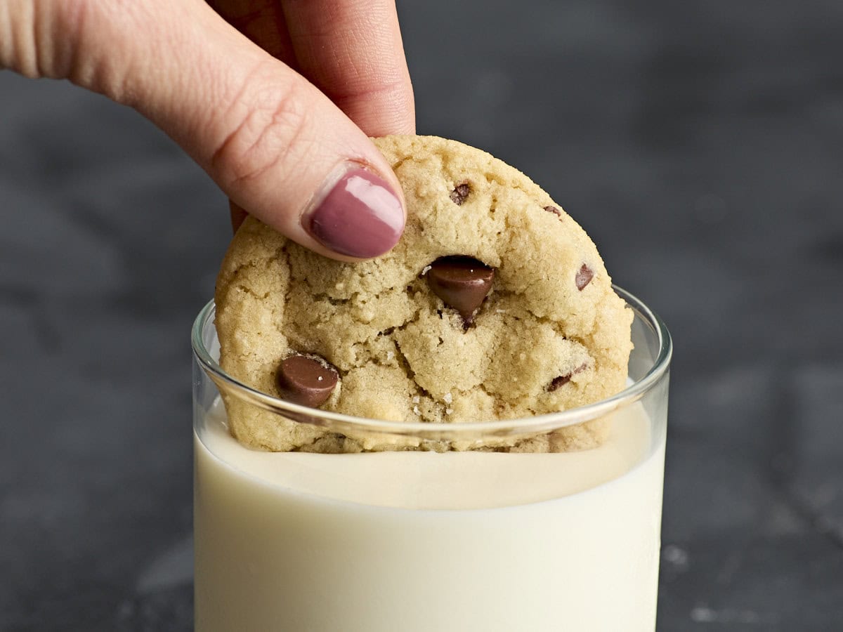 A hand dunking a chocolate chips cookie into a glass of milk.