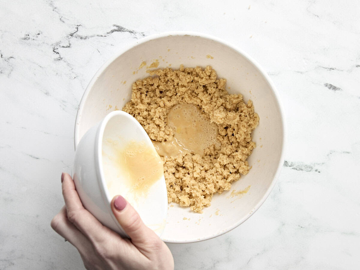 Beaten egg being poured into a bowl with creamed butter and sugar.