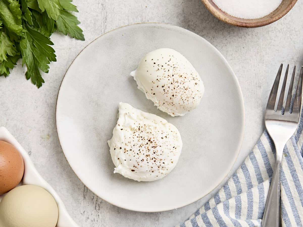 Overhead view of two poached eggs on a plate, topped with salt and black pepper.