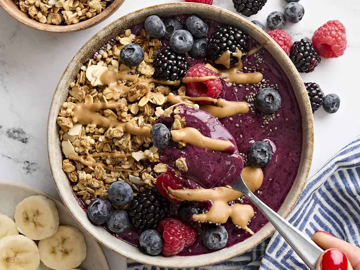 Overhead view of a spoon taking a spoonful from a smoothie bowl, which is topped with granola, chia seeds, berries, and peanut butter.