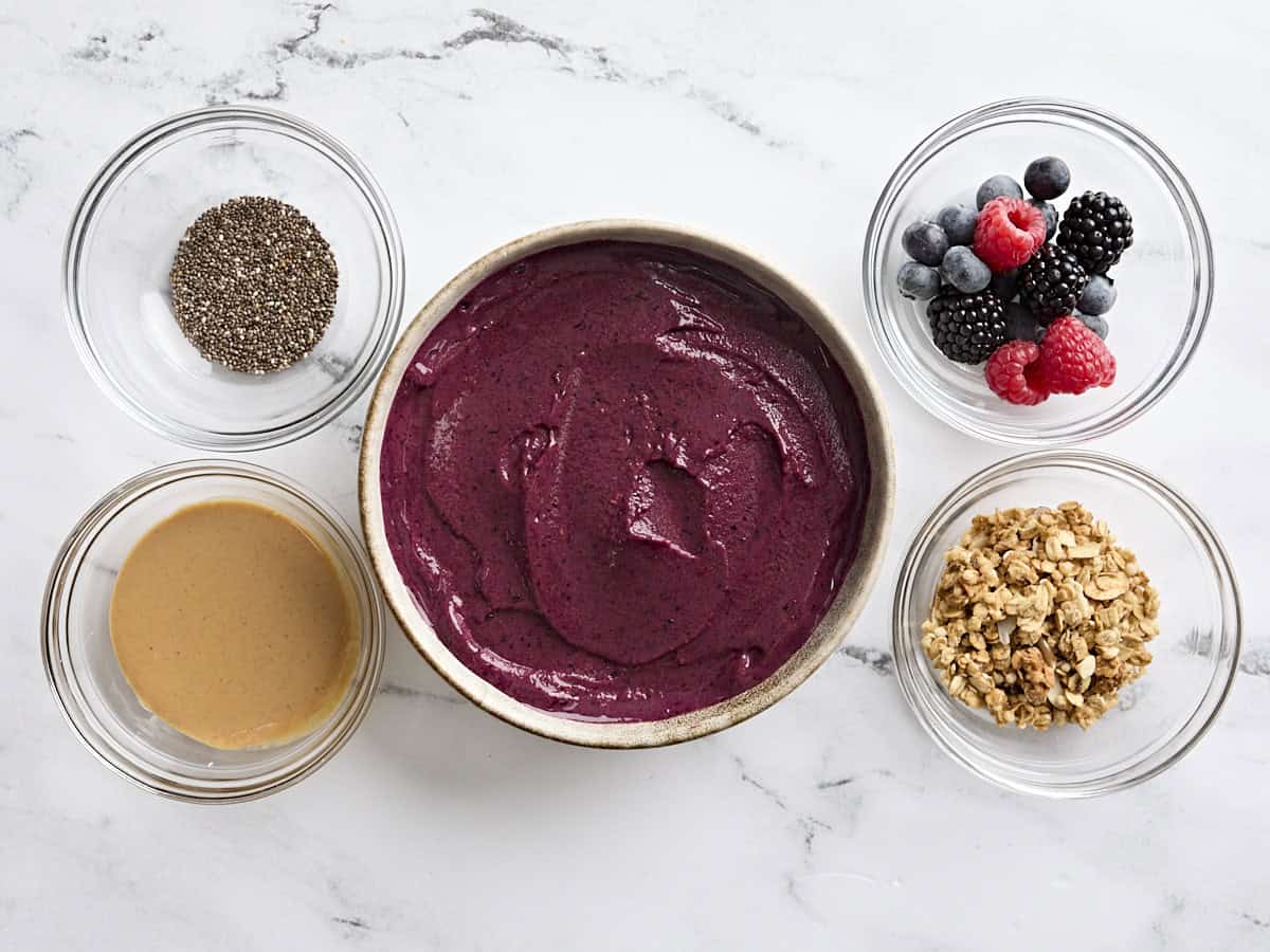 A smoothie bowl surrounded by various toppings in smaller bowls, including berries, granola, peanut butter, and chia seeds.