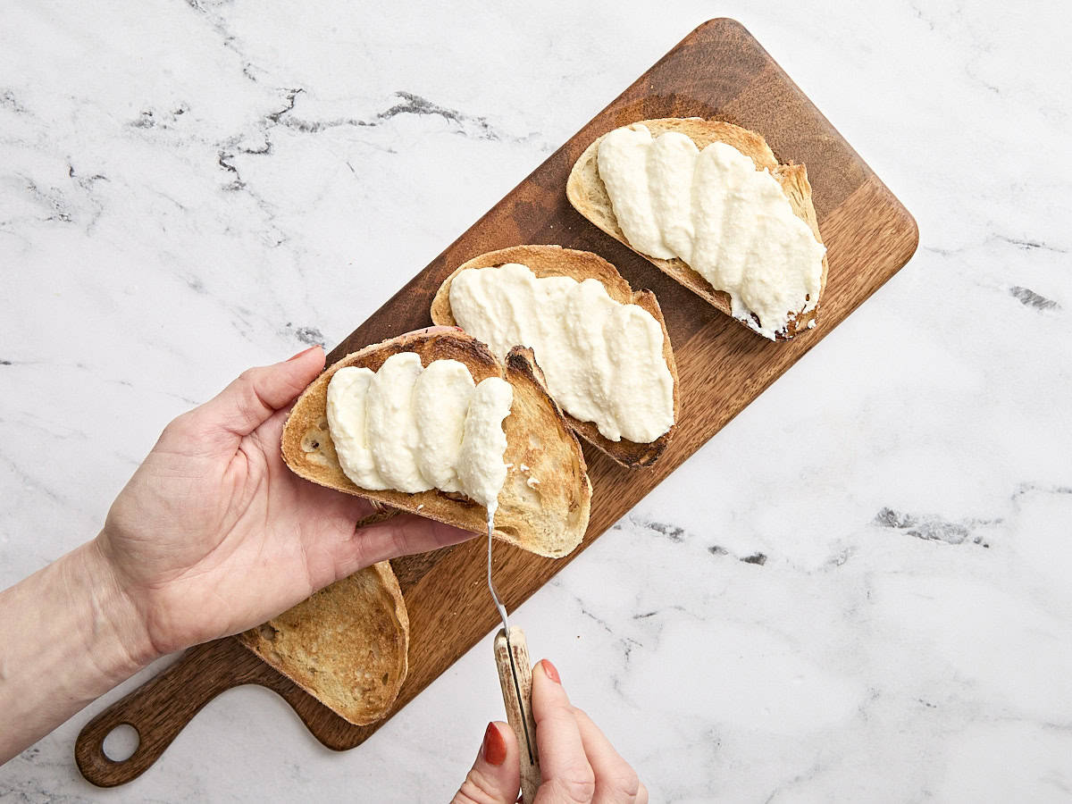 A hand spreading whipped ricotta on slices of sourdough toast.