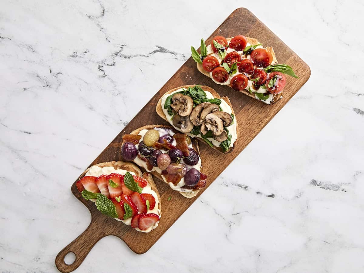 Ricotta toast 4 ways on a wooden serving platter, one topped with strawberries and mint, the next with roasted grapes and bacon, another with blistered tomatoes and basil, and the last with mushrooms and wilted spinach.