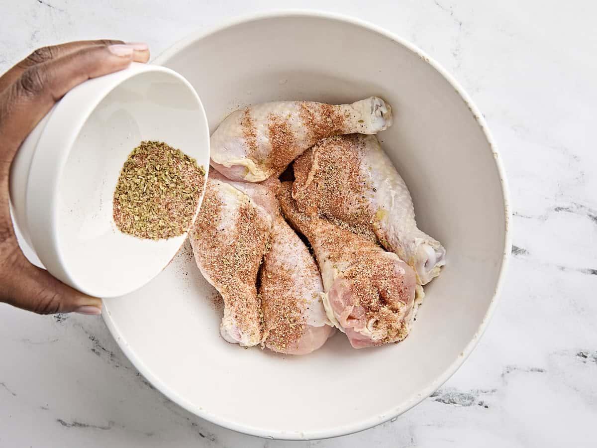 Seasoning being added to chicken drumsticks in a bowl.